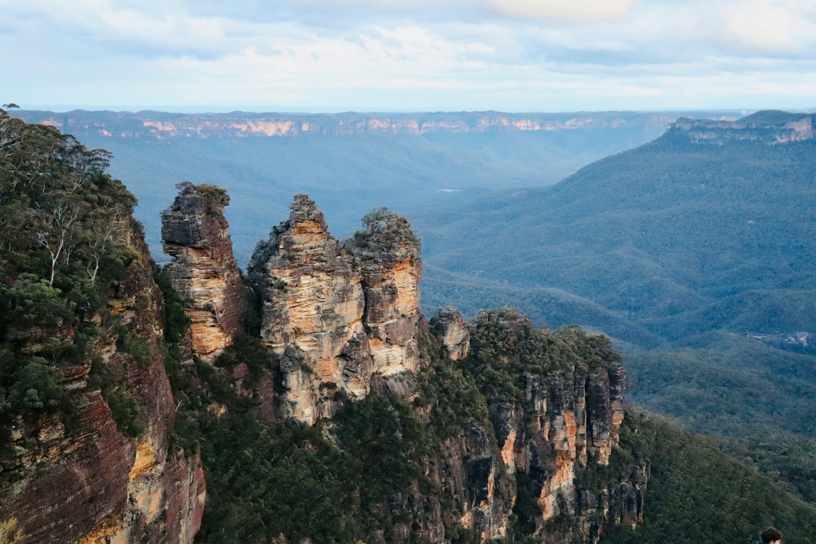 The Three Sisters rock formation glowing amber above a wide, blue-hazed eucalyptus valley in the Blue Mountains near Katoomba.