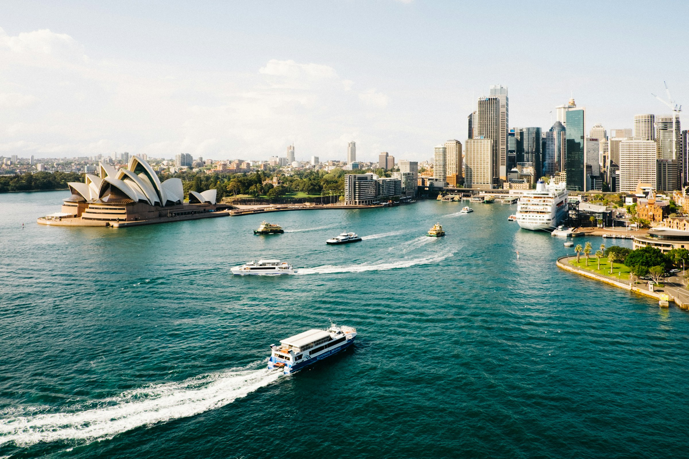 Sydney Harbour at midday, the Opera House sails on the left, the CBD skyline on the right, and green-and-yellow ferries carving white wakes across the blue water.