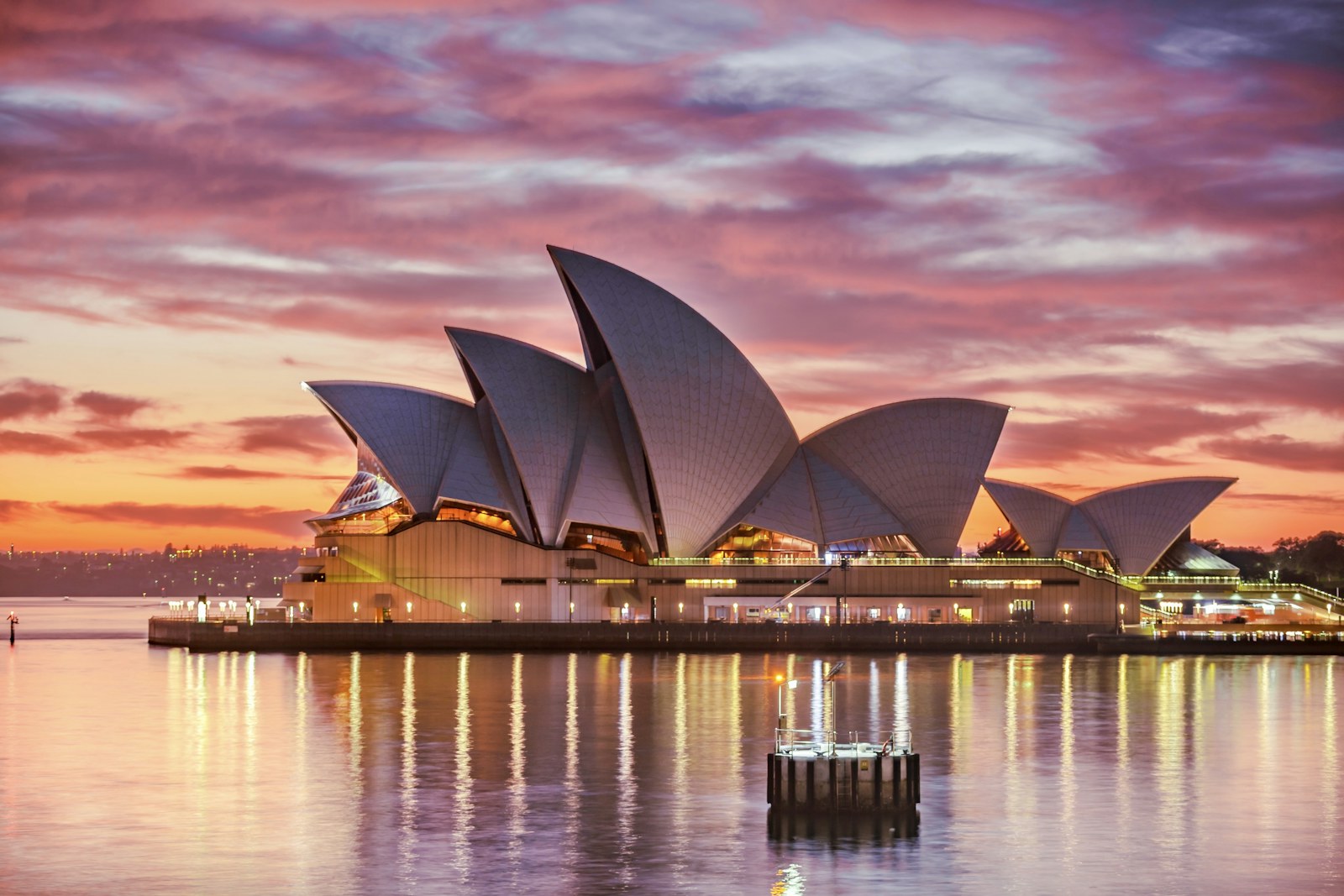 The Sydney Opera House sails under a vivid pink and orange sunset, warm lights glowing along the harbour promenade.